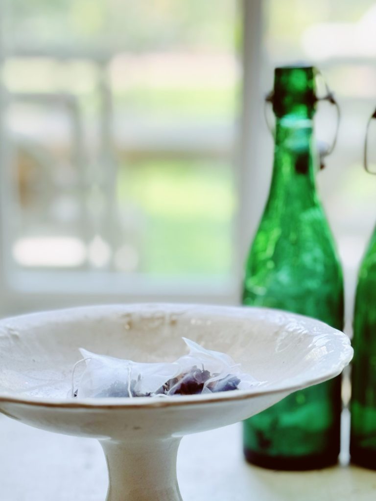 A close-up of a aromatic tea bags in a white ceramic bowl with two green glass bottles in the background, near a window.