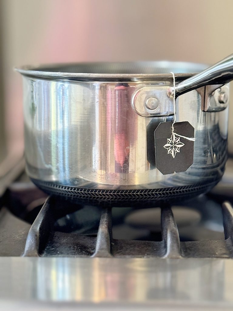 A stainless steel pot on a gas stove burner with a tea bag hanging from the handle.
