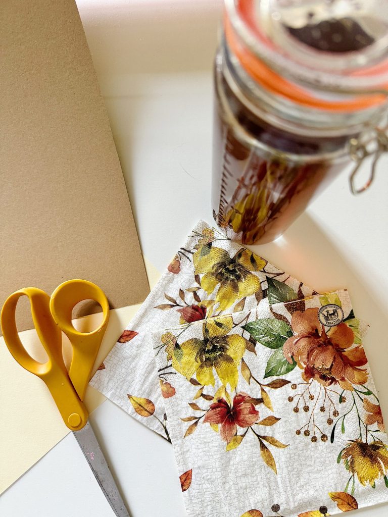 A desk with craft supplies including yellow scissors, floral fabric, and a jar of tea, with a blank paper sheet in the background.