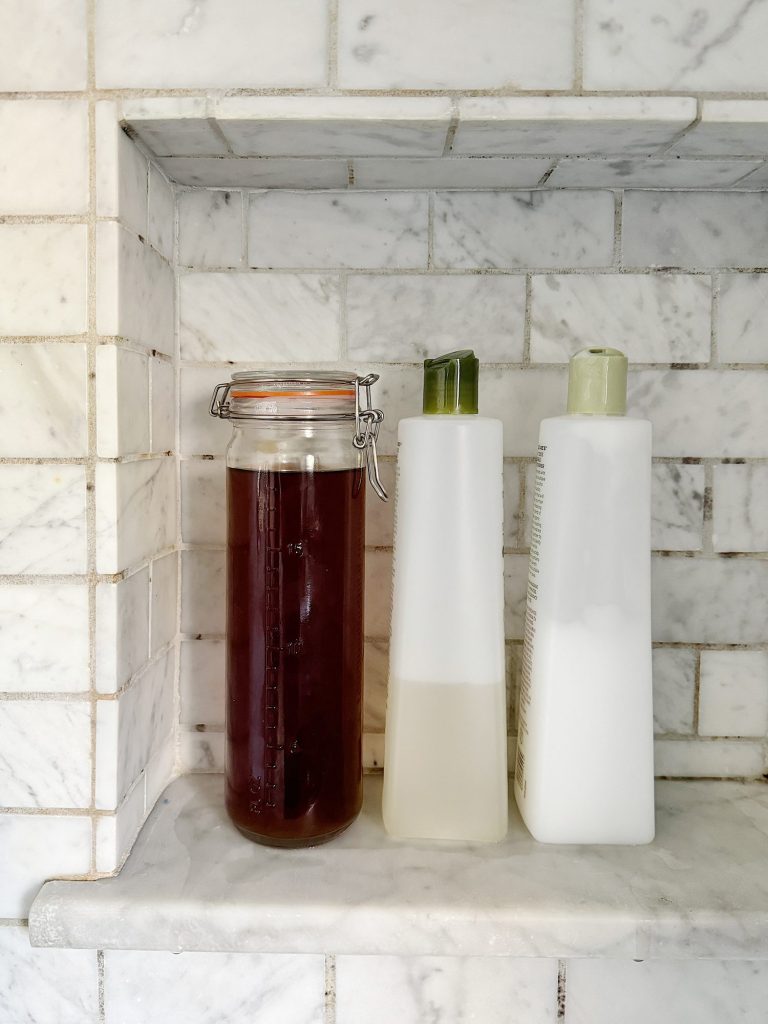 Three containers—a glass jar of tea and two bottles with pumps—sit on a marble shelf in a tiled niche of a shower.
