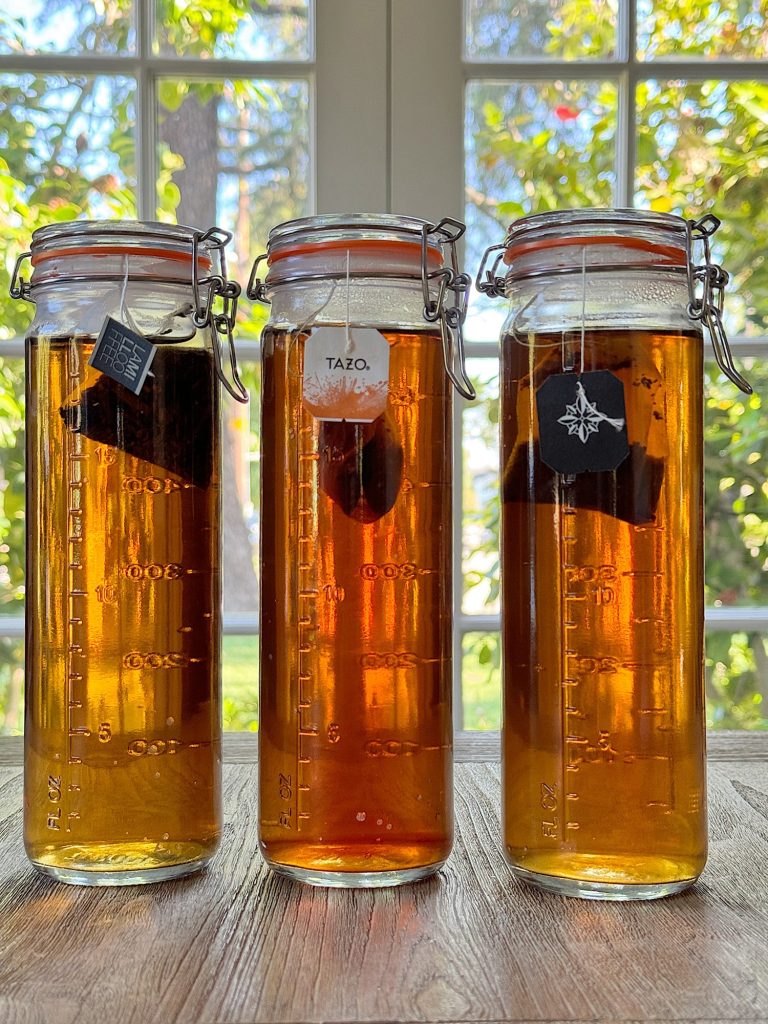 Three glass jars containing different shades of iced tea, with tea bags visible, set on a wooden surface in front of a window with garden view.