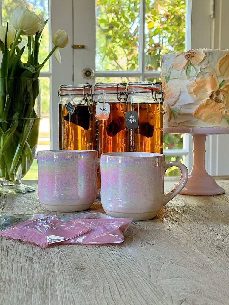 Three glass jars containing different shades of iced tea, with tea bags visible, set on a wooden surface with two mugs, dlowers, and a cake in front of a window with garden view.