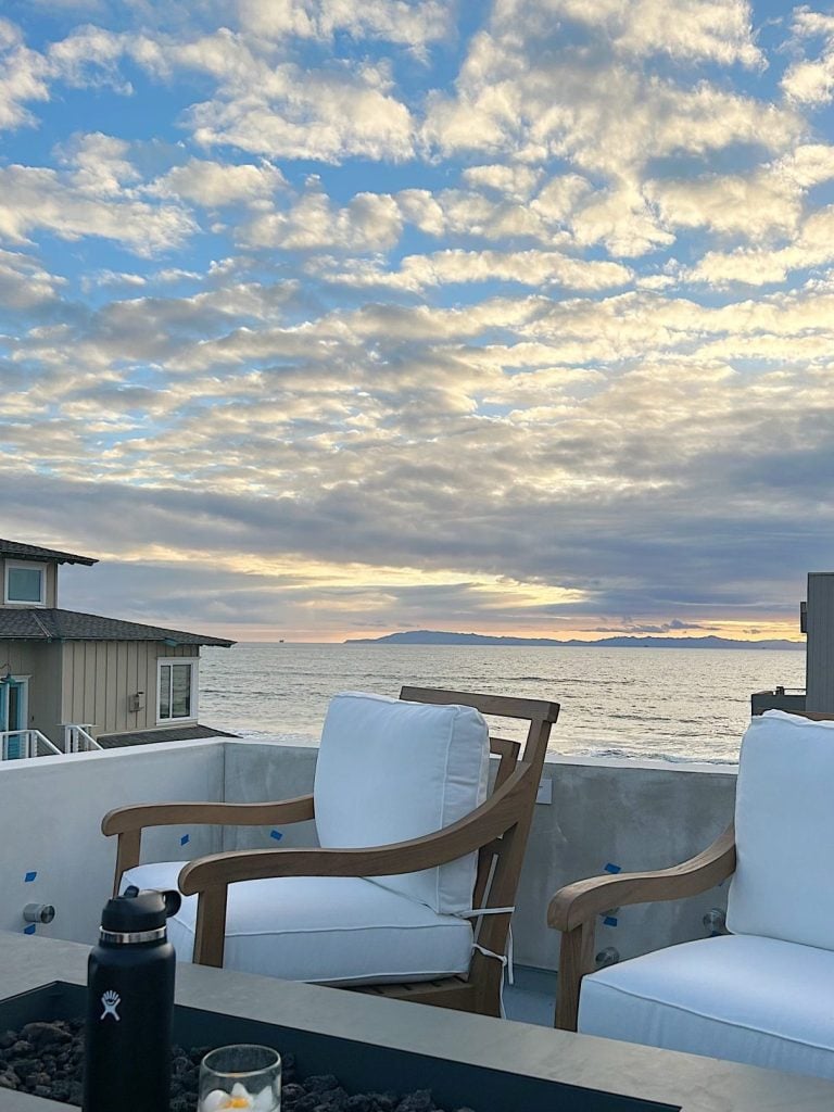 Two white cushioned chairs on a patio overlook the ocean at sunset. A black water bottle and a glass candle sit on the table in front of the chairs. Patchy clouds fill the sky above the horizon.