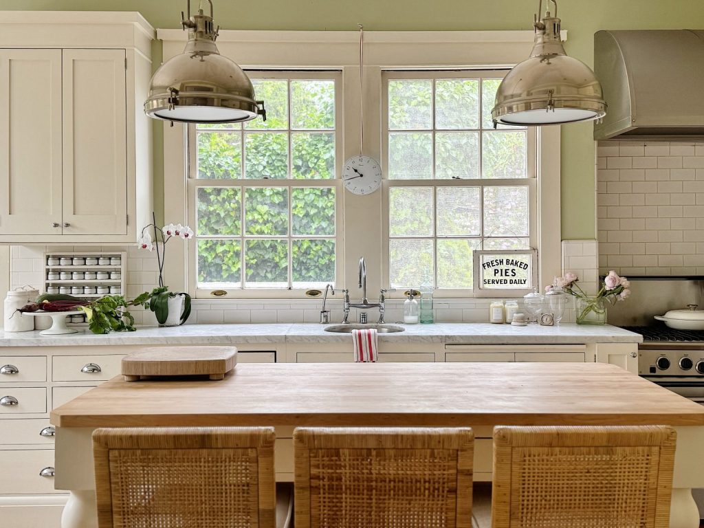 A kitchen with a wooden island, three rattan stools, white cabinets, metal pendant lights, and a large window above the sink. A sign reads "Fresh Baked Pies Served Daily.