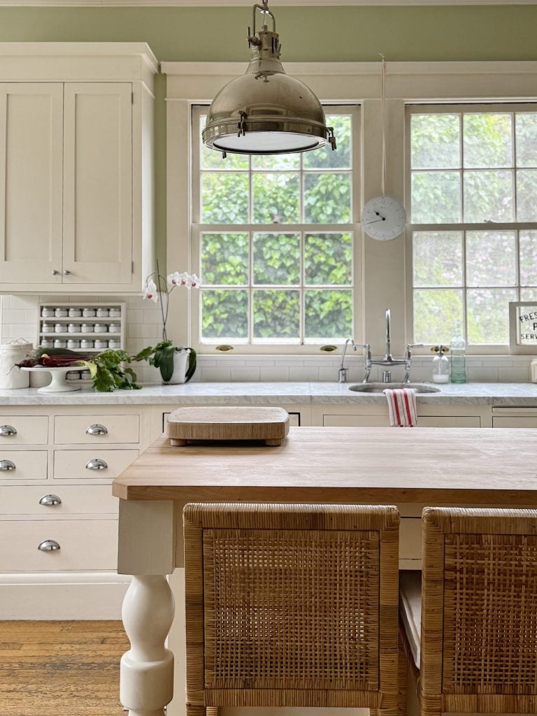 A bright, airy kitchen with a wooden island and two wicker chairs, white cabinets, a large hanging lamp, and a sink under a window with lush green foliage outside.