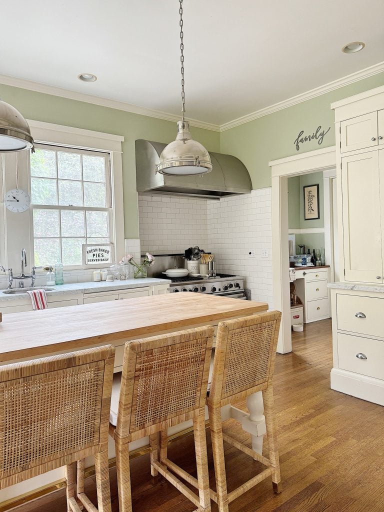 A kitchen with light green walls, a large window, three rattan bar stools at the island, white cabinetry, pendant lights, and a door leading to an adjacent room. The word "family" is on the wall.