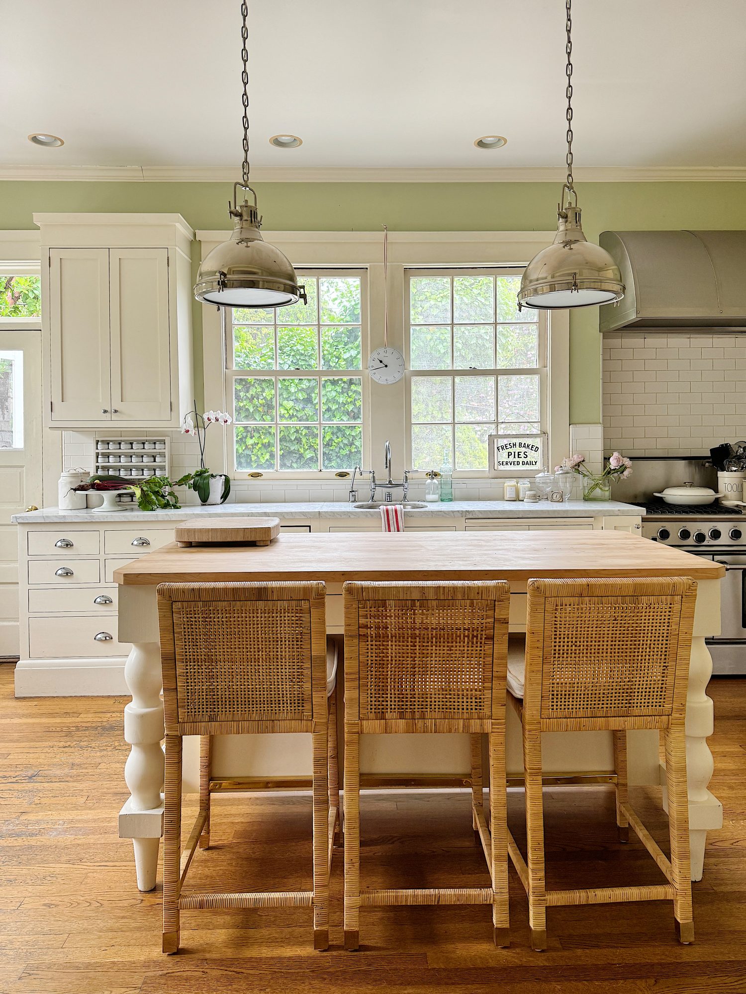 A bright kitchen with white cabinets, a wooden island, and three woven chairs. Two pendant lights hang above the island, and a large window provides natural light. The floor is wooden.
