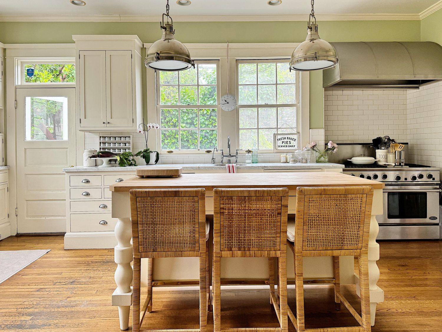 A bright kitchen featuring a large island with woven chairs, a stainless steel stove, two windows, and hanging pendant lights. There are white cabinets and a door to the side.