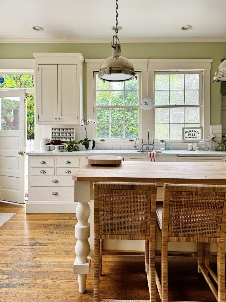 A bright kitchen with white cabinets, a wooden island with two wicker bar stools, pendant lights, and large windows overlooking greenery. The back door is slightly open.