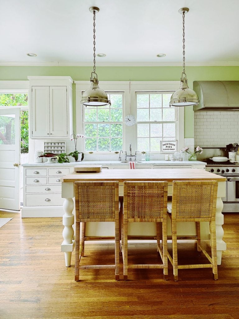 A bright kitchen features a white island with two wicker barstools, pendant lights, stainless steel appliances, white cabinetry, and large windows.