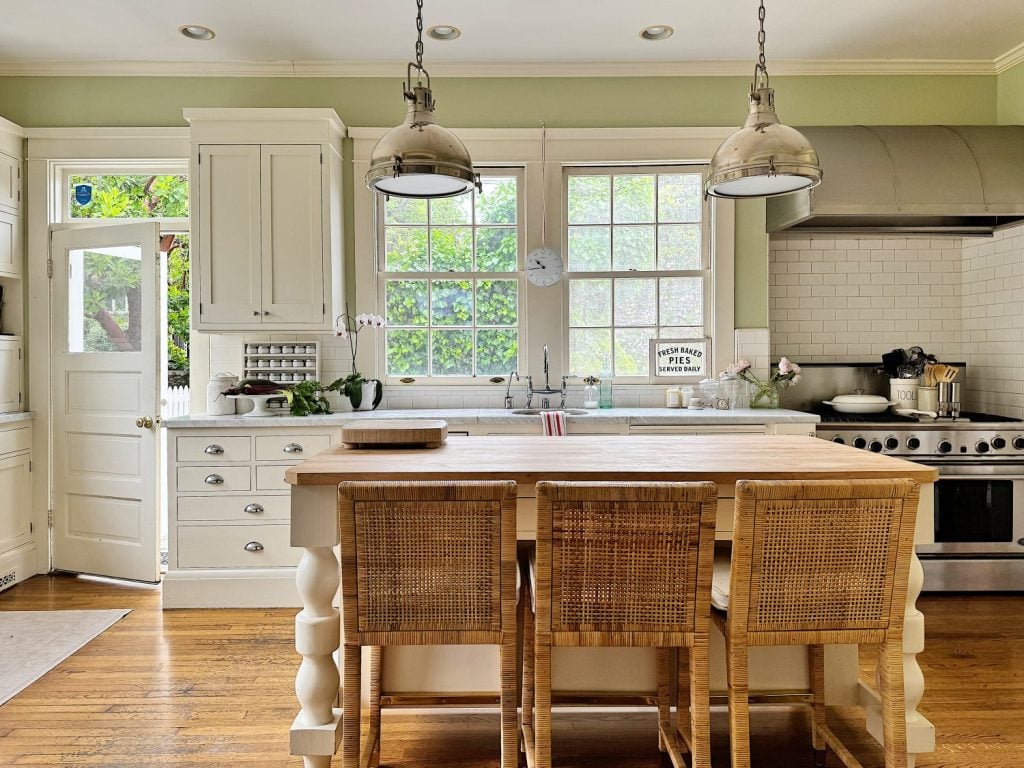 A bright kitchen with white cabinets, a central wooden island with three wicker chairs, stainless steel appliances, and large windows letting in natural light. Hanging lights illuminate the space.