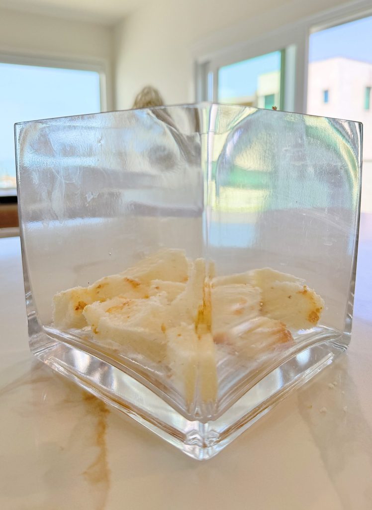 Glass container with a few pieces of torn bread placed on a light-colored marble surface, set in a bright, sunlit room.