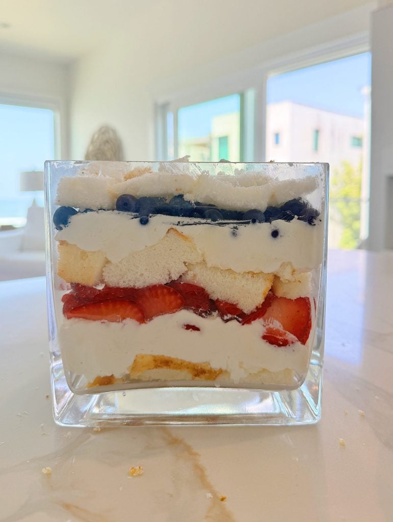 A glass container on a table filled with layers of whipped cream, blueberries, sponge cake, and sliced strawberries, with windows and blurred buildings in the background.