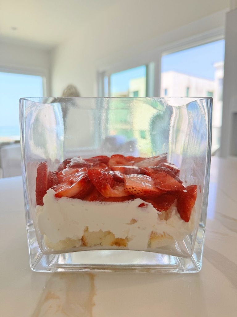 Glass container filled with layered dessert featuring a base of ladyfingers, a middle layer of creamy filling, and a top layer of sliced strawberries, set on a white countertop.