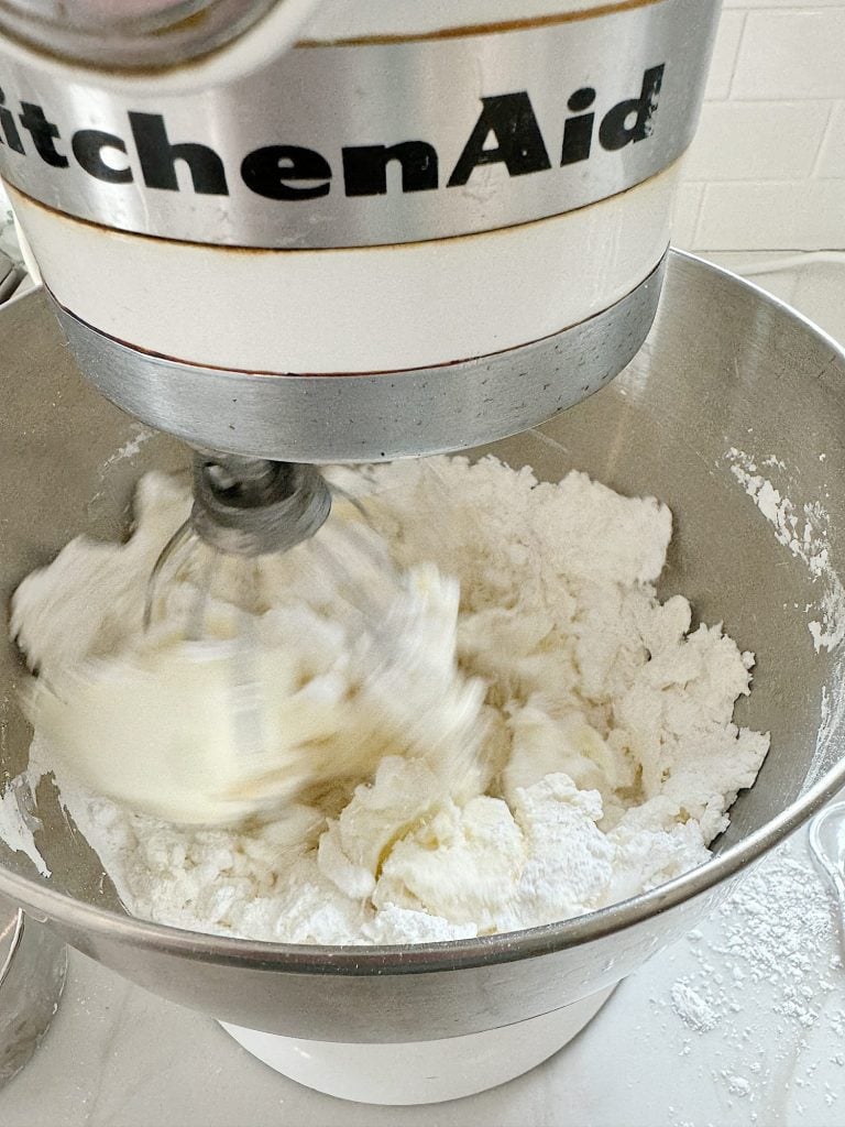A KitchenAid stand mixer blending white icing in a stainless steel bowl. The mixture appears fluffy and is being mixed with a wire whisk attachment.
