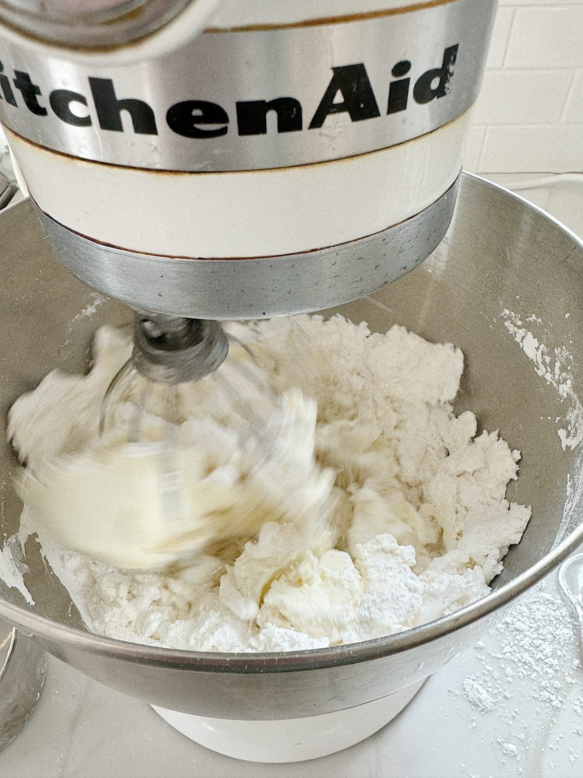 A KitchenAid stand mixer blending white icing in a stainless steel bowl. The mixture appears fluffy and is being mixed with a wire whisk attachment.