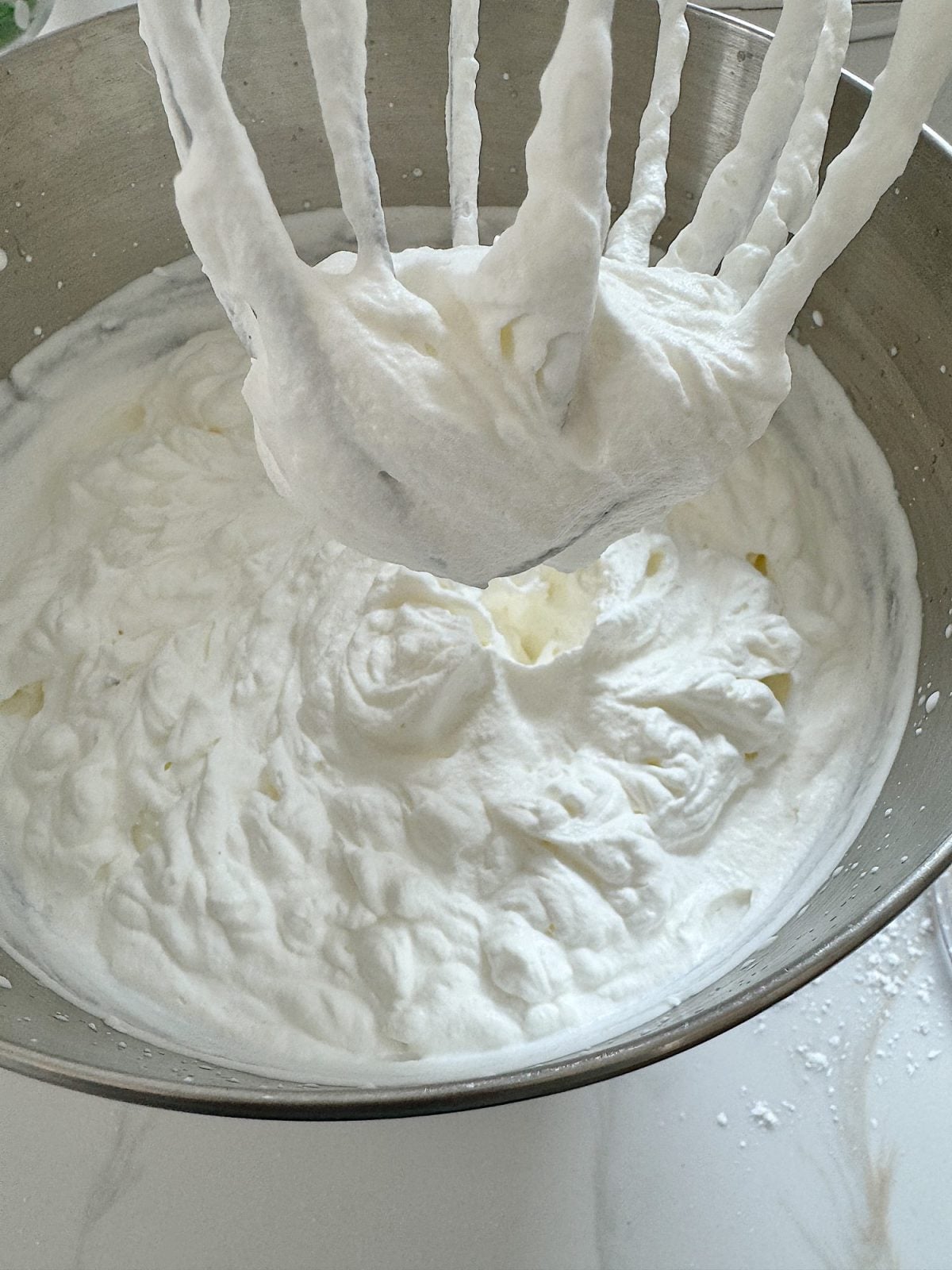 A metal mixing bowl filled with whipped cream, with a whisk attachment covered in whipped cream hovering above the bowl.
.