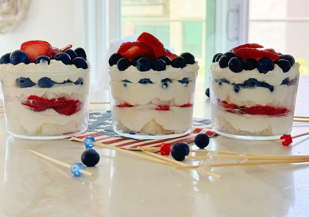 Three layered desserts in glass cups with blueberries, strawberries, and cream, arranged on a marble countertop with scattered blueberries and skewers in the foreground.
