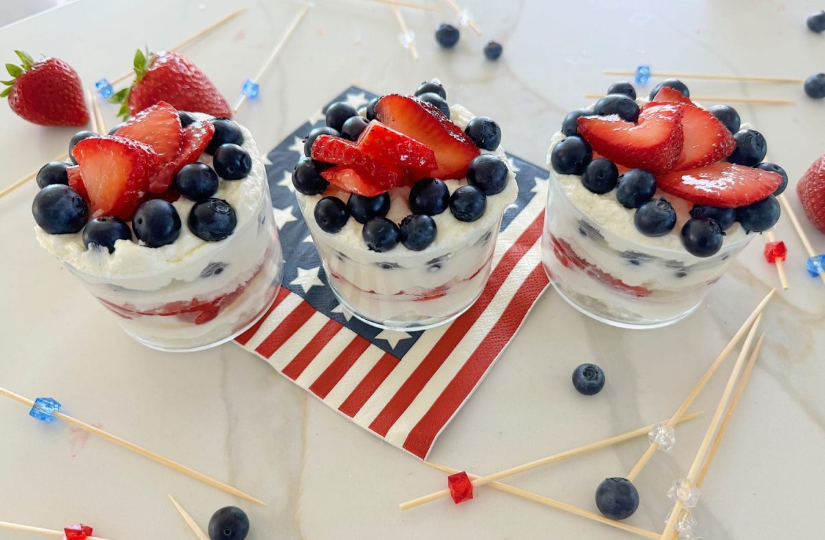 Three layered desserts in glass cups with blueberries, strawberries, and cream, arranged on a marble countertop with scattered blueberries and skewers in the foreground.