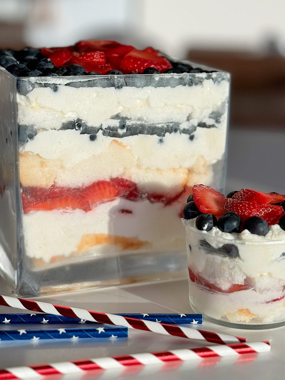 A layered dessert in a large and small glass dish with blueberries and strawberry slices on top, cream, and cake pieces in between, designed to resemble a trifle.