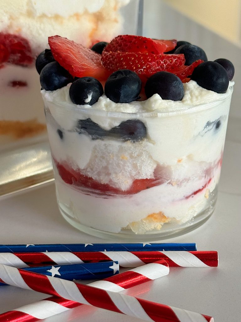 A layered dessert in a large and small glass dish with blueberries and strawberry slices on top, cream, and cake pieces in between, designed to resemble a trifle.