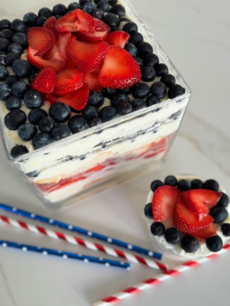 A layered dessert in a large and small glass dish with blueberries and strawberry slices on top, cream, and cake pieces in between, designed to resemble a trifle.
