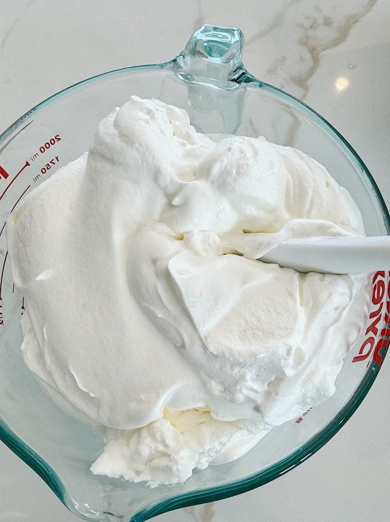 A glass measuring cup filled with whipped cream and a white spatula partially submerged in the mixture.