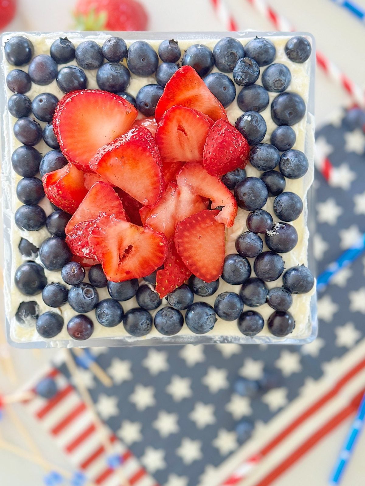 Square cake topped with a heart-shaped arrangement of sliced strawberries and surrounded by blueberries, placed on a star-patterned napkin. Red-and-white striped straws are scattered in the background.