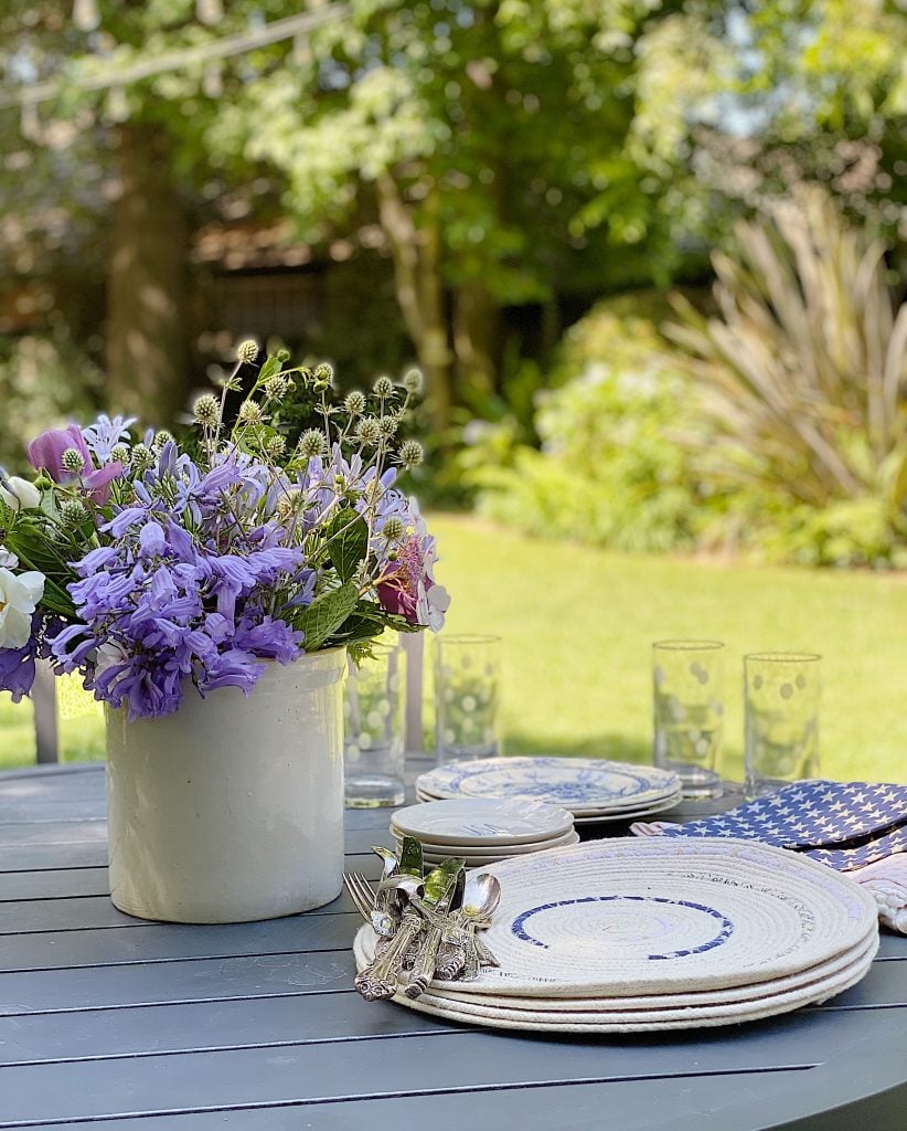Outdoor table set with plates, glasses, and a white vase containing purple and white flowers. The scene is in a garden with greenery in the background.