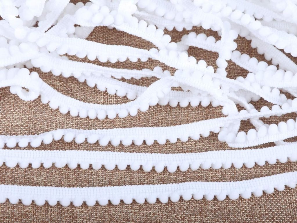 White pom pom trim ribbon is spread out on a textured brown fabric background, perfect for adding a touch of patriotic decor to your next craft or celebration.
