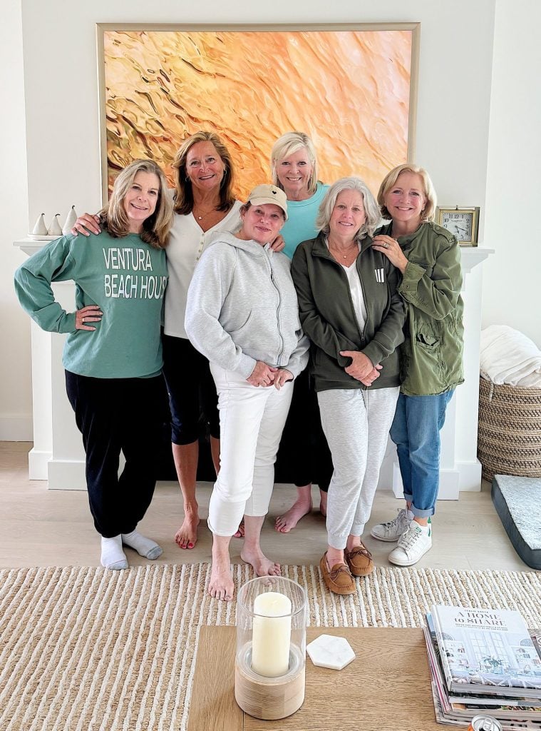 Six women standing together in a living room, posing for a photo. They are casually dressed and smiling, with a large painting on the wall behind them and a coffee table in the foreground.