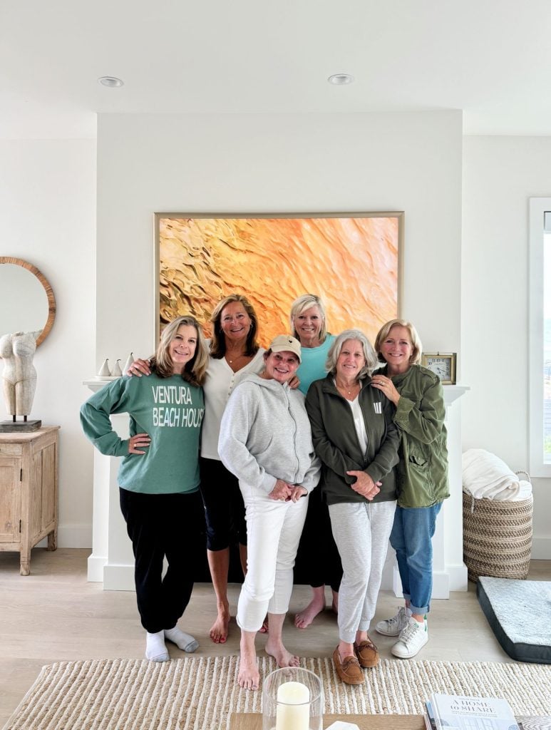 Six women standing close together in a living room, all smiling at the camera. A large painting and a statue are in the background. The room is bright and modern.