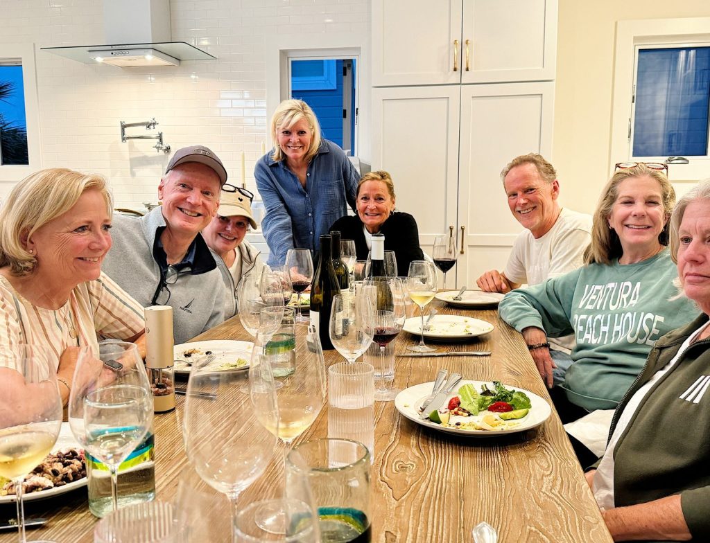 A group of eight people smiling and seated around a dining table with plates of food and glasses of wine in a well-lit kitchen.