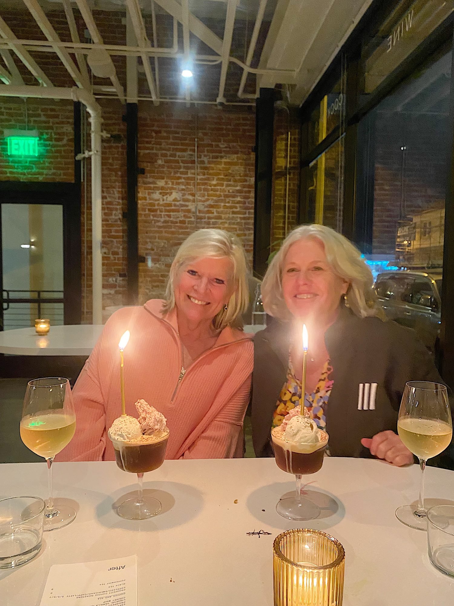 Two women smiling while sitting at a table with three glasses of wine and two dessert cups with lit candles in a dimly lit restaurant.