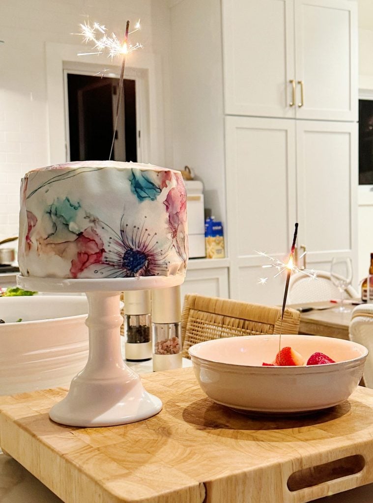 A cake decorated with floral patterns and sparklers on a white stand next to a bowl of strawberries with a sparkler, set on a wooden surface in a kitchen.