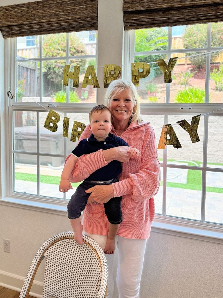 A woman in a pink sweater holds a baby in a room decorated with a "Happy Birthday" banner hanging from a window.