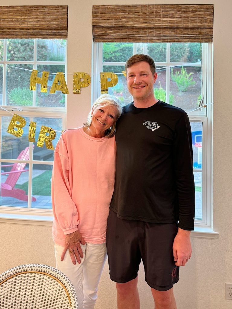 An older woman in a pink sweater and a tall man in a black shirt stand side by side in front of a window with "HAPPY BIRTHDAY" decorations.
