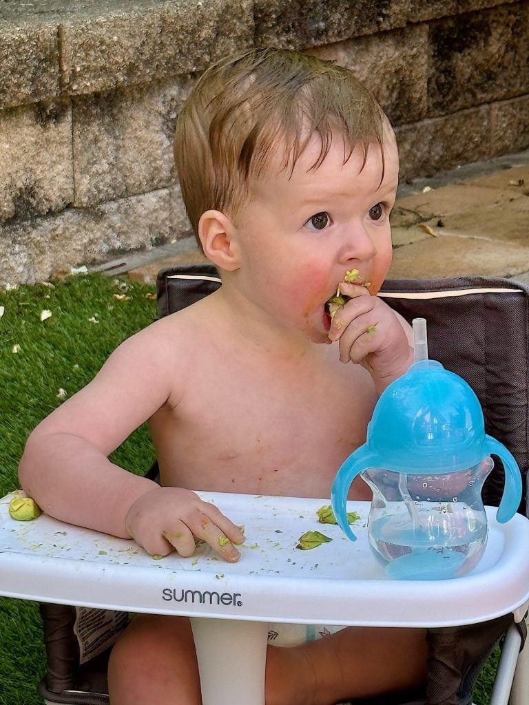 A baby sits in a high chair outdoors, eating with one hand and holding food in the other. A blue sippy cup filled with water is placed on the tray in front of the baby.