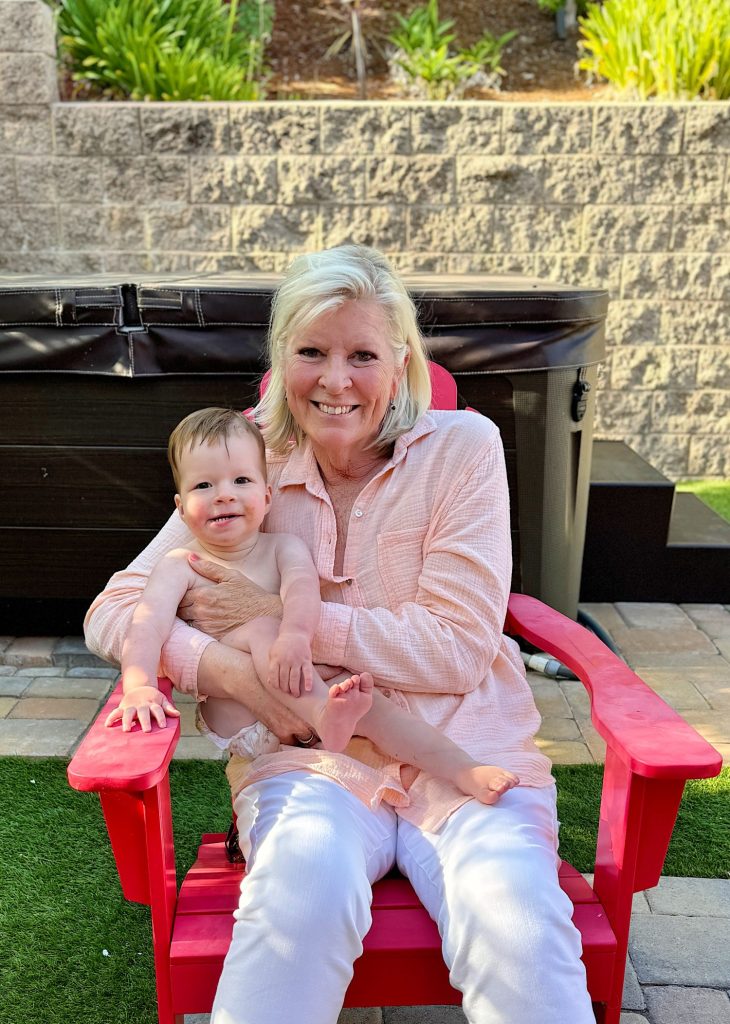 An older woman with shoulder-length blonde hair sits on a red chair outdoors, holding a smiling baby on her lap. Both are dressed in light-colored clothing. A hot tub is visible in the background.