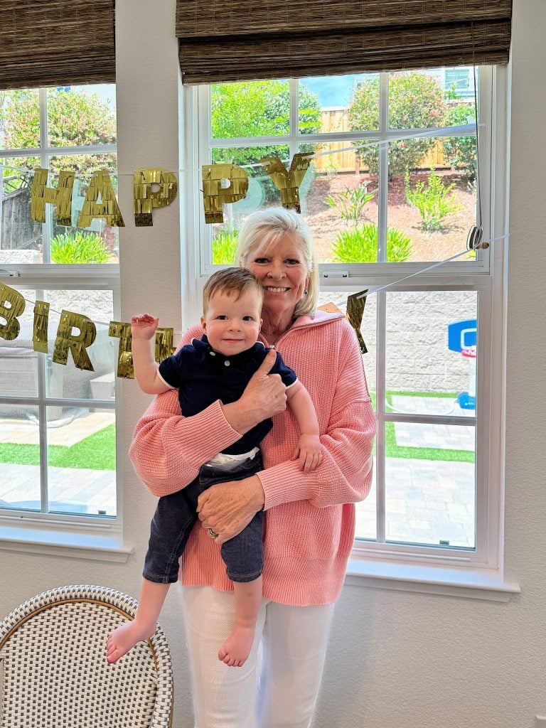 An older woman holds a young child in her arms in front of a window with a "Happy Birthday" banner hanging behind them. Both are smiling.