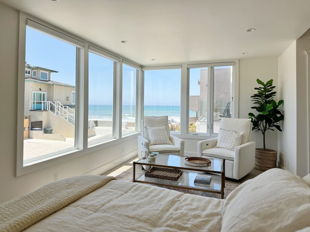 A modern bedroom with large windows overlooking a beach, featuring a bed, two white chairs, a glass coffee table, and a potted plant.