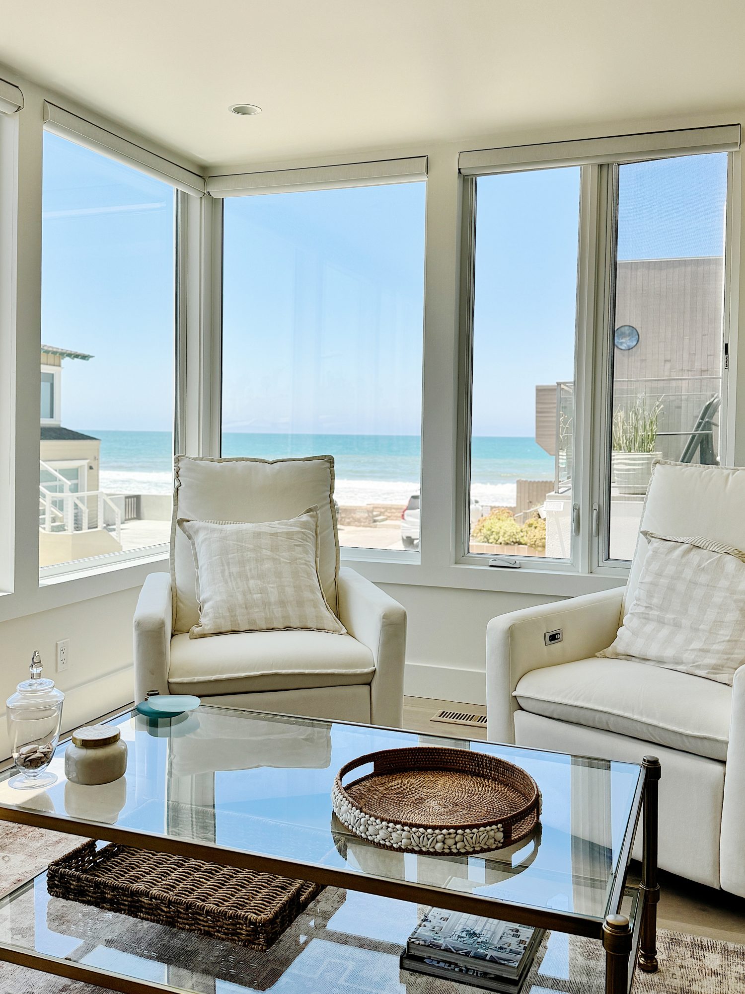 A bright room with large windows offering an ocean view, featuring two white armchairs, a transparent coffee table, and a coastal-themed tray.