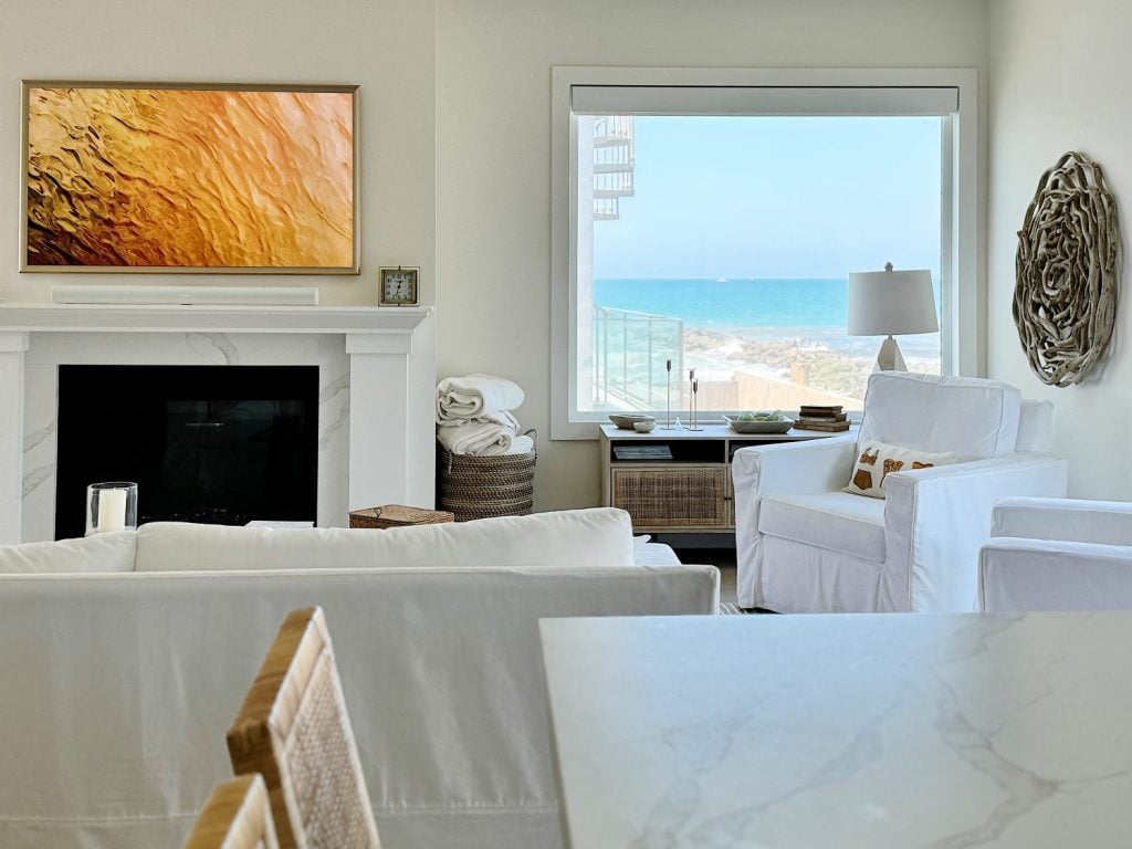 A bright, airy living room with white furniture, a wall-mounted TV showing abstract art, a large window revealing an ocean view, and minimalist decor.