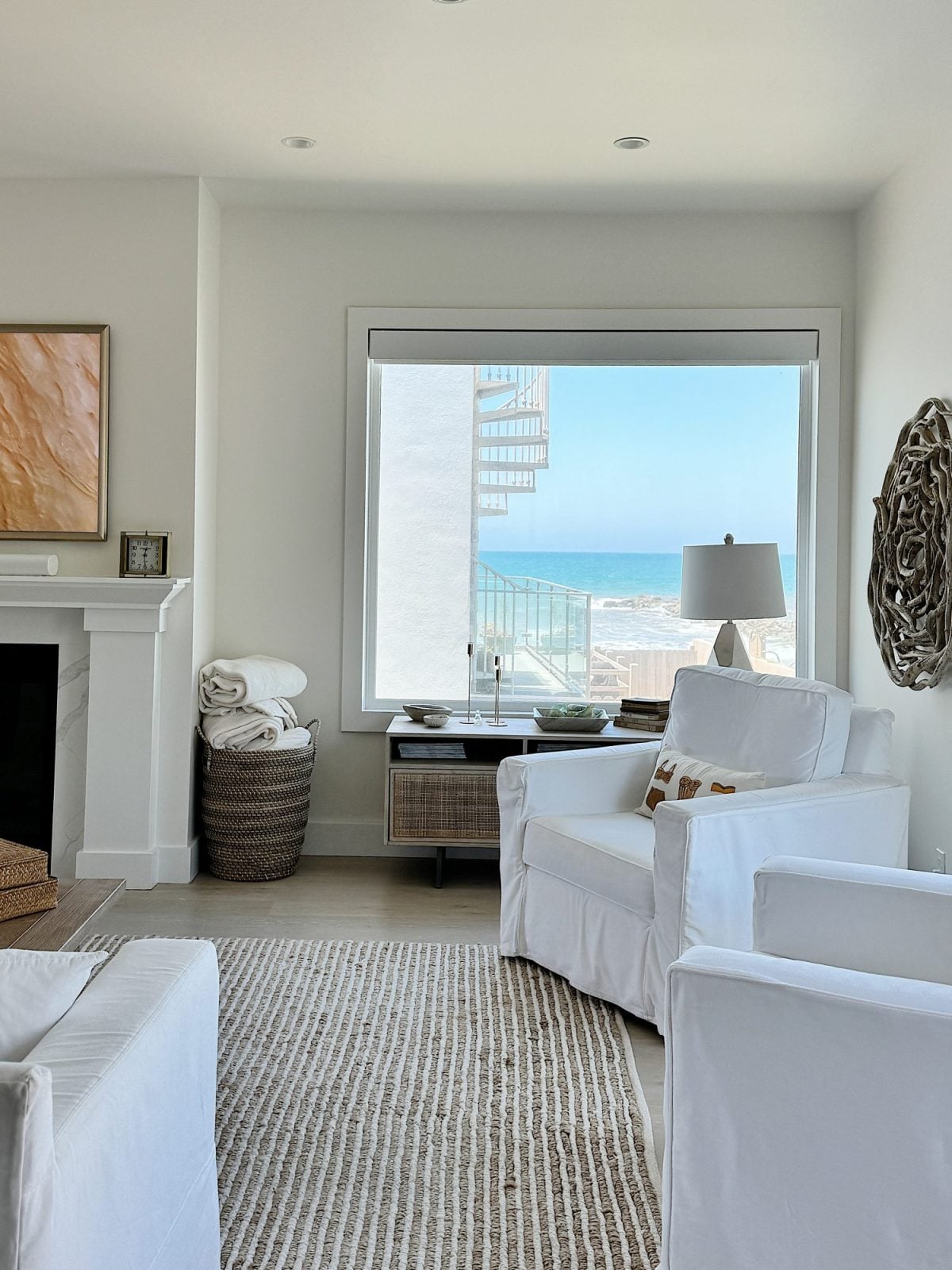 A bright beachside living room with white furniture, a large window showing a sea view, a spiral staircase outside, and coastal decor including a woven basket and wall art.