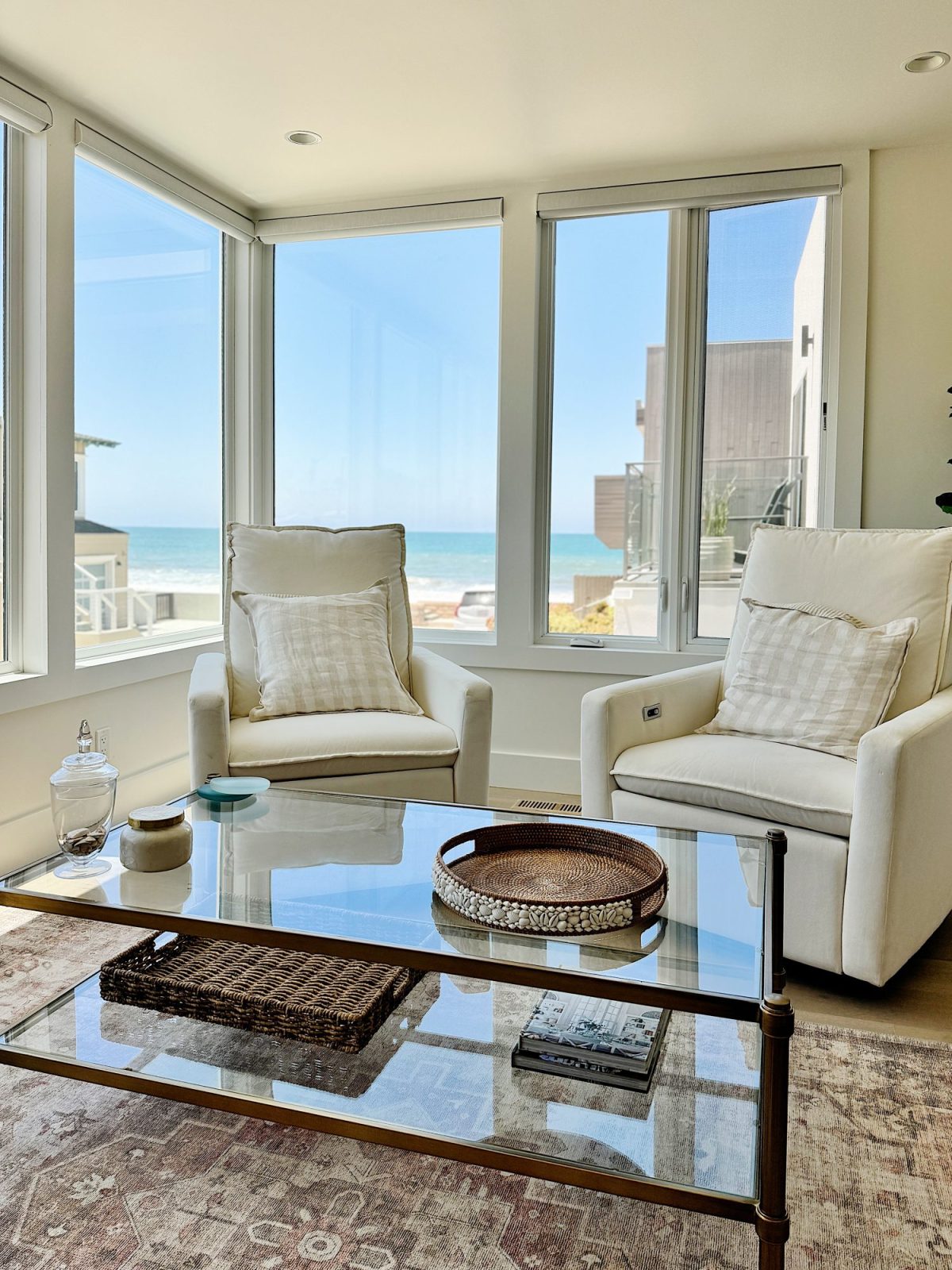A sunlit living room with two armchairs and a glass coffee table. Large windows provide a view of the beach and ocean. The coffee table has decorative items, including a tray and a small basket.