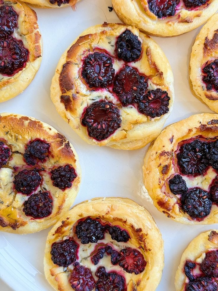 Small pastries topped with blackberries are arranged on a white surface.