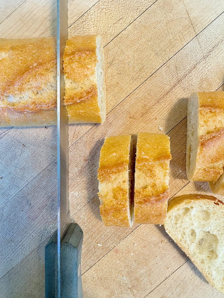 A knife slicing a baguette on a wooden cutting board. Several slices are already cut and placed near the whole piece.