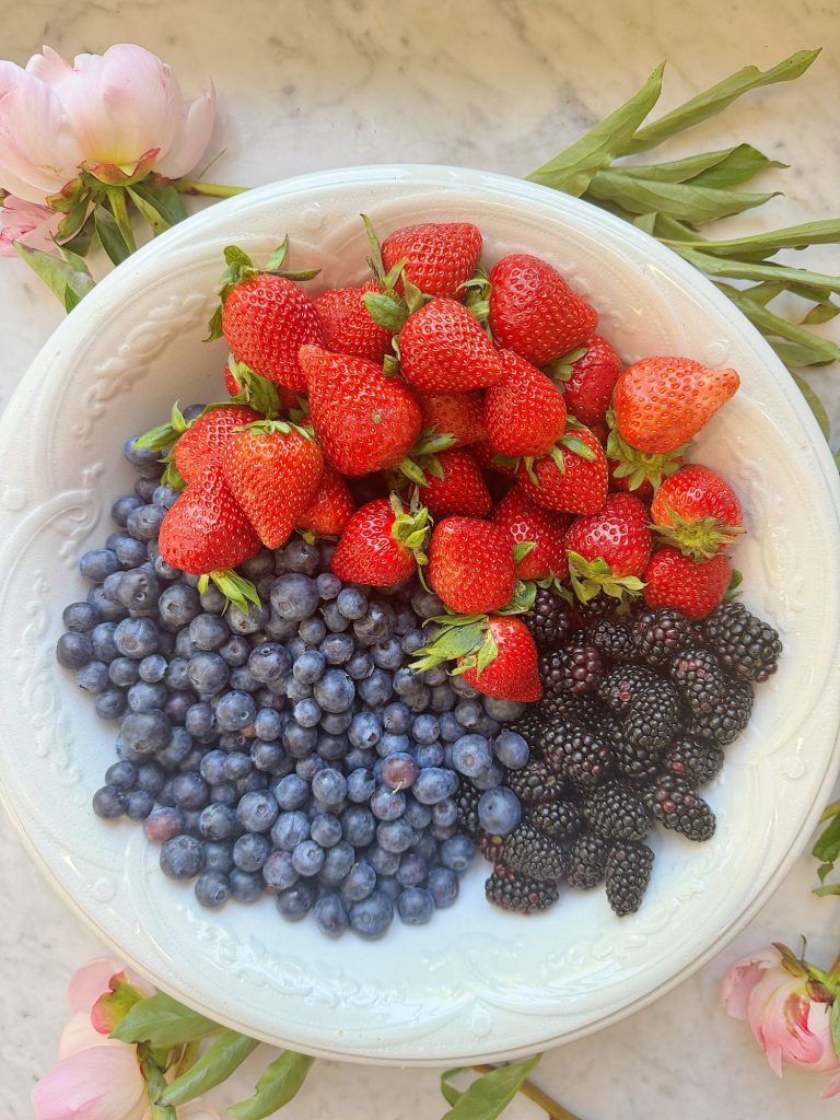 A white plate contains fresh strawberries, blueberries, and blackberries. Pink flowers and green stems are visible around the plate.