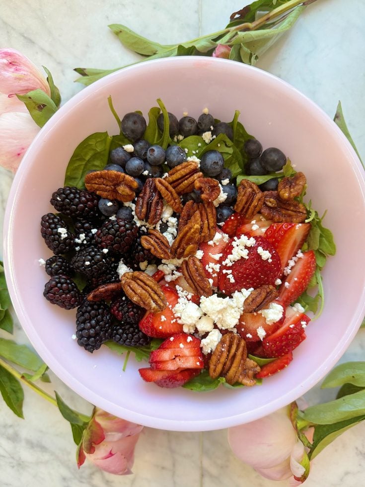A bowl of salad with blackberries, blueberries, strawberries, spinach, pecans, and crumbled cheese on a light-colored surface with petals around the bowl.