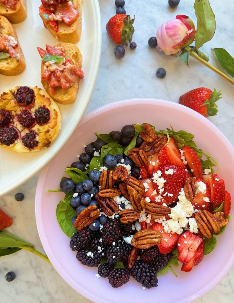 A bowl of salad with strawberries, blueberries, blackberries, pecans, and feta cheese beside a plate with bruschetta and berry-topped tarts, all set on a marble surface with scattered berries and a flower.